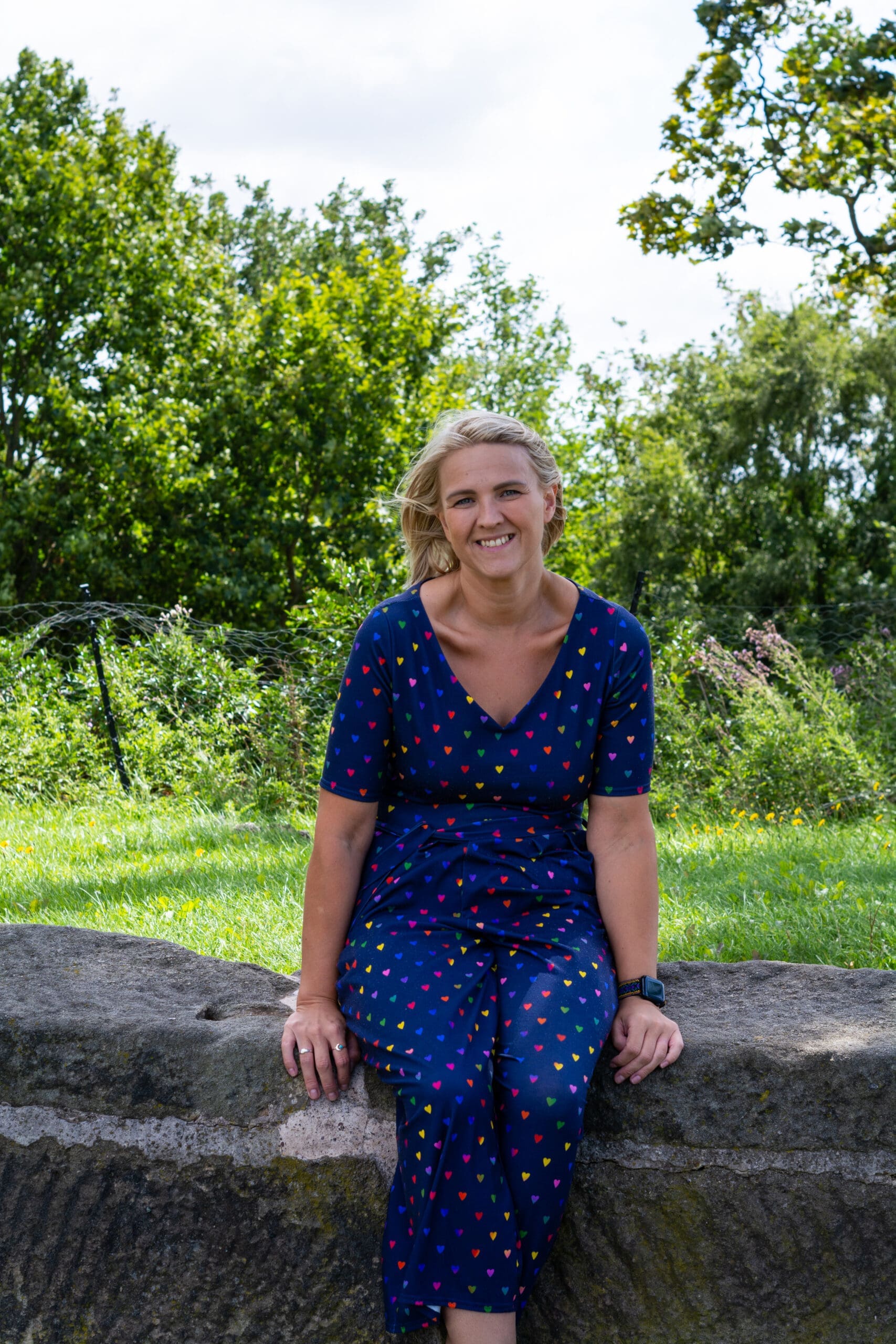 Melissa in a blue, heart-patterned jumpsuit sits on a stone ledge against a backdrop of lush green trees, smiling warmly on a sunny day.